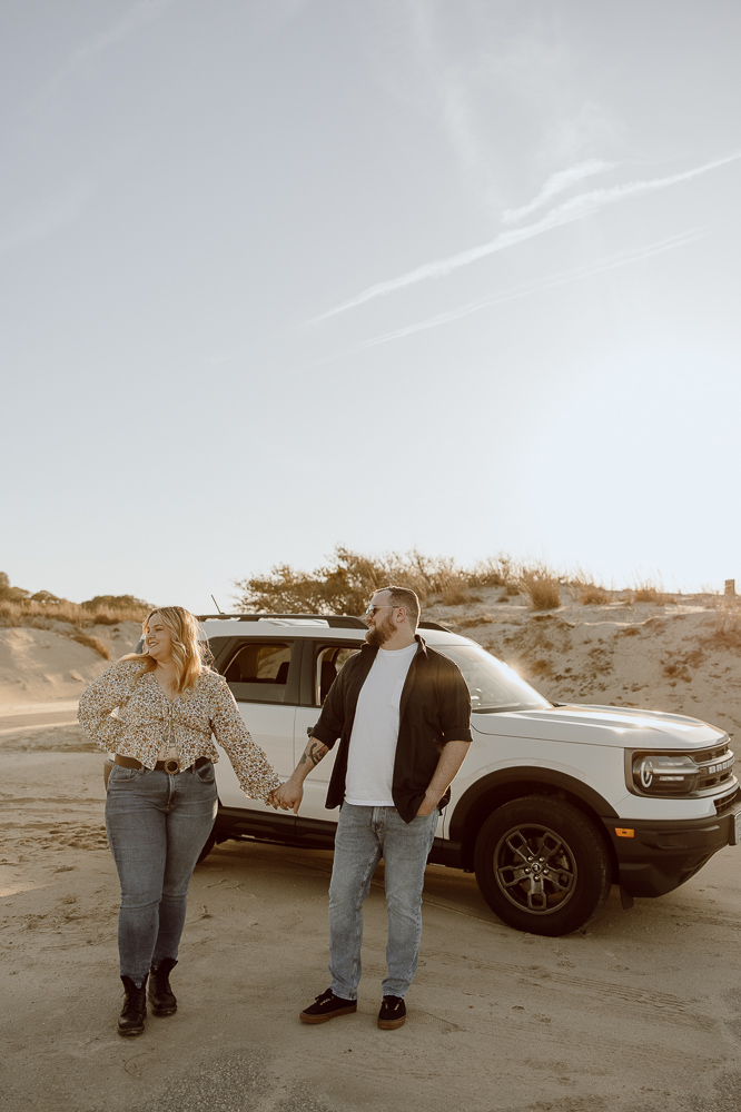 couple holding hands and looks to their side as they stand in front of a car.