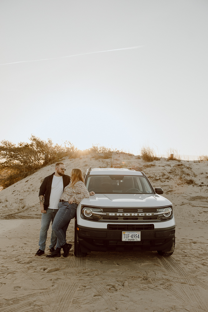 Couple leans and props an arm onto their car while staring at each other.