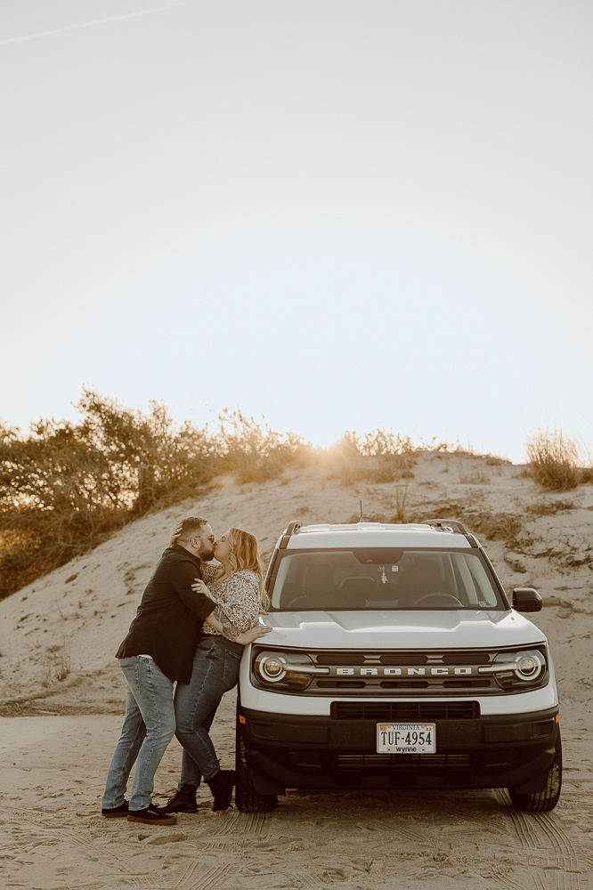 Couple shares a kiss while leaned against the side of their car.