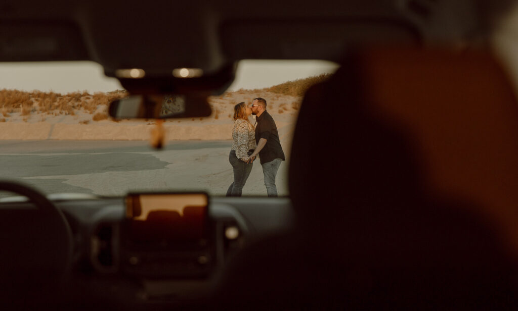 couple stands outside of car and shares a kiss, seen through the windshield.