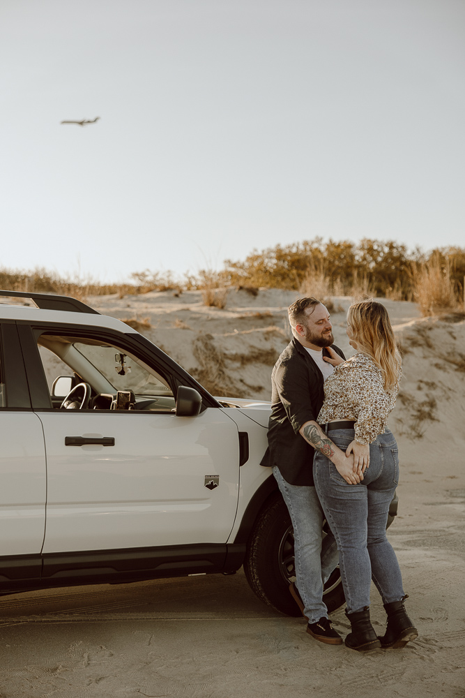 couple leans onto their car and hold each other close as a plane flys over.