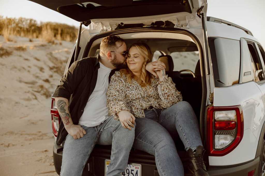 Couple shares a kiss while sitting in the open trunk of a car during sunset.