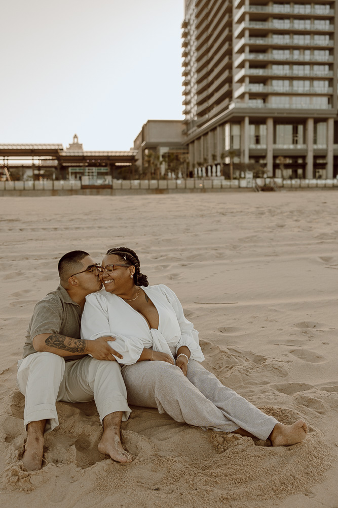 Man kisses his partner on the cheek as they sit next to each other on the beach.