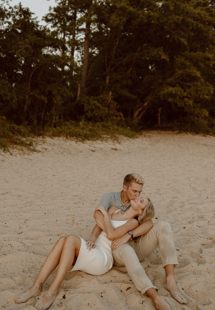 Woman leans onto her partner as they sit on the beach and he kisses her forehead.