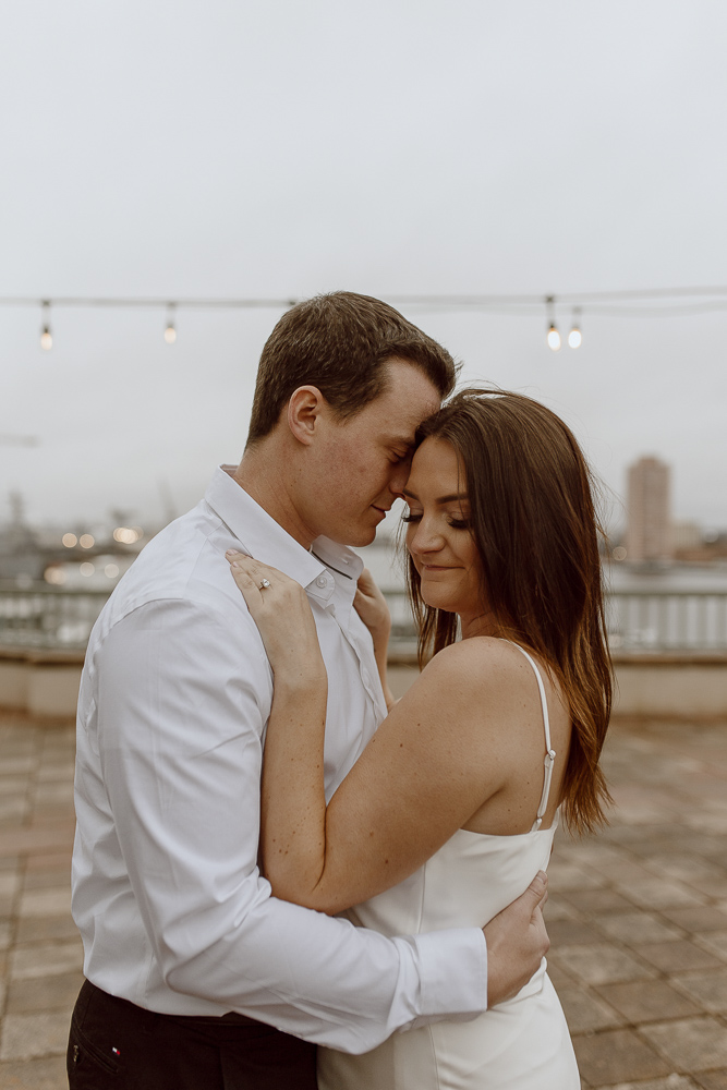 Couple stands close and smiles with Norfolk Virginia skyline behind them.