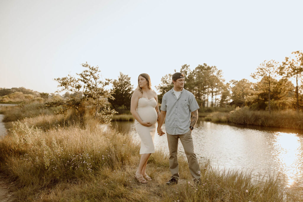 Expecting mom and her husband hold hands and stand while looking in opposite directions.