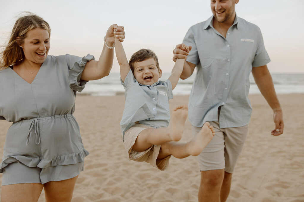 On the beach, parents swing their toodler son in the air as he smiles in joy.