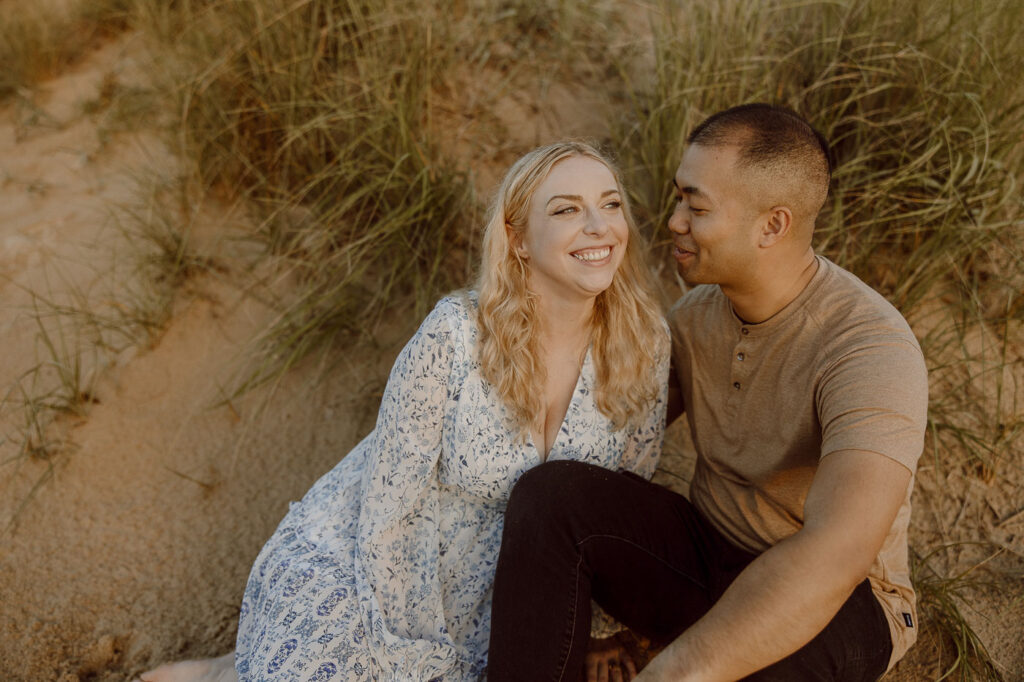 Couple smiles at each other while sitting on sand dunes in Virginia Beach. 