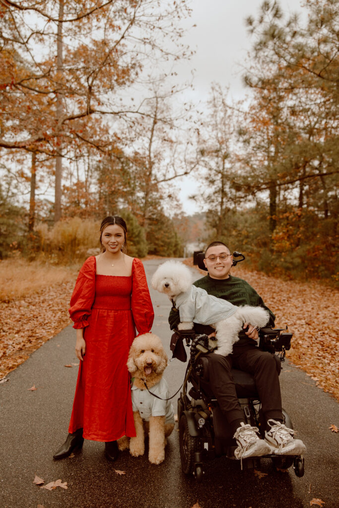 couple and their dogs stand and smile while surrounded by fall folliage. 