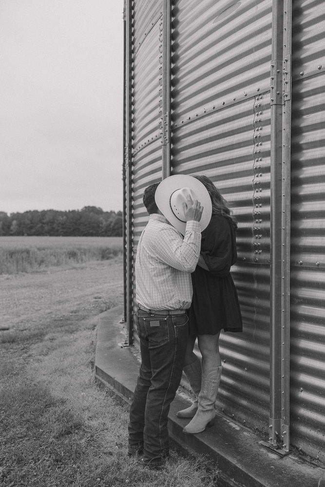 Couple leans against grain pin on a farm while blocking the view of a kiss with his cowboy hat.