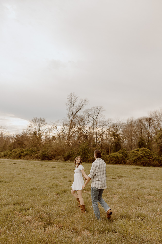 Engaged couple walking through field while holding hands.