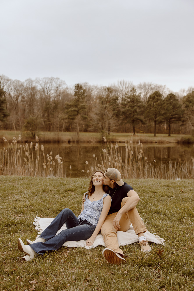 Couple sits on a blanket near a pond and shares laughs.