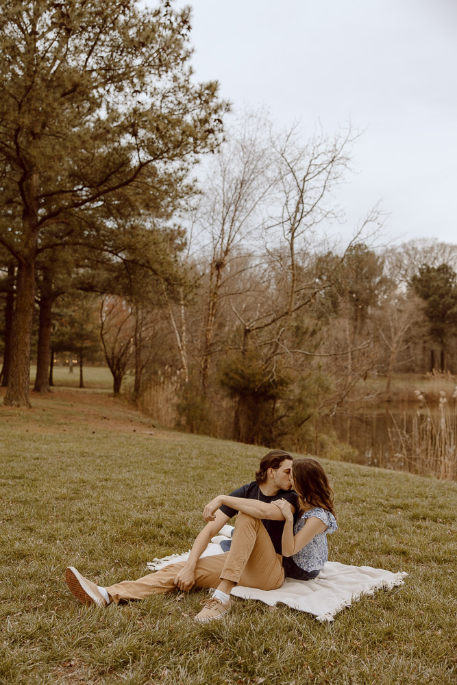 Couple shares a kiss while sitting on blanket near a pond.