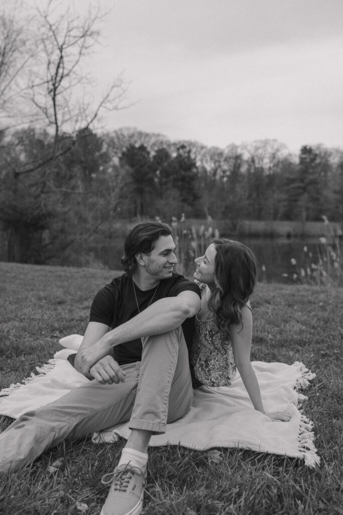 Couple smiling at each other while sitting on a blanket in front of a pond.