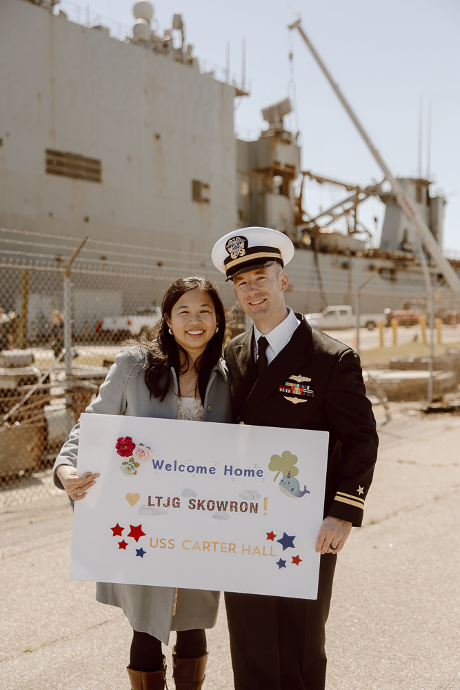 A navy sailor and his wife smiling in from of a navy ship.