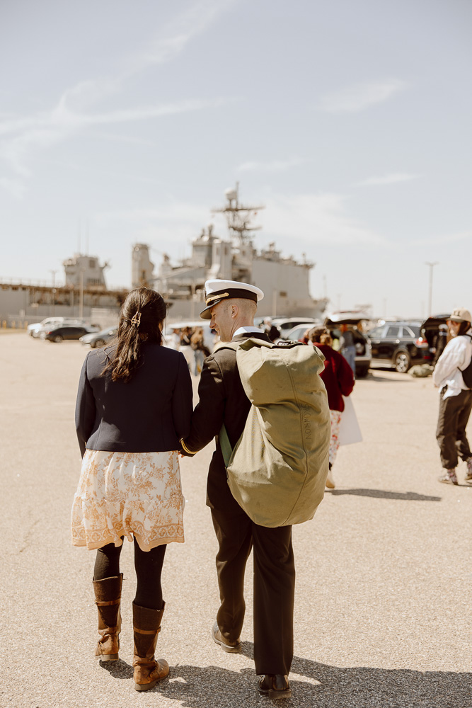 A military sailor and spouse walking away from a Navy ship while holding hands.