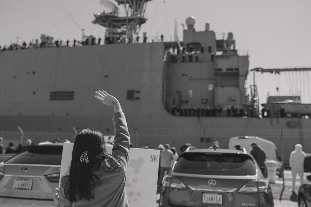 A military wife waves to her husband who is standing on a Navy ship.