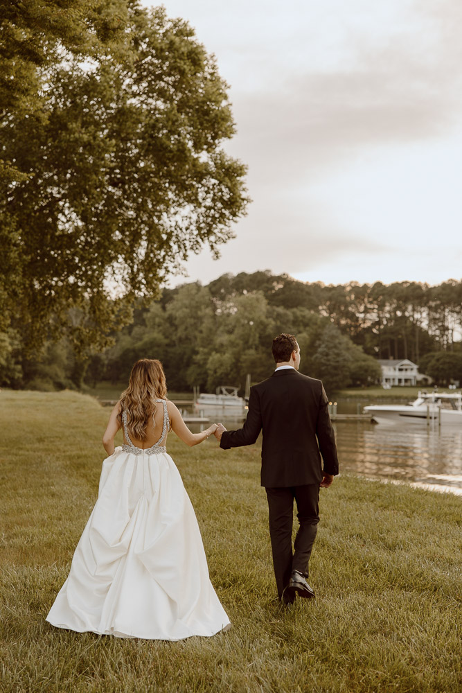 A couple in wedding attire hold hands as they walk through the grass near a boat dock.