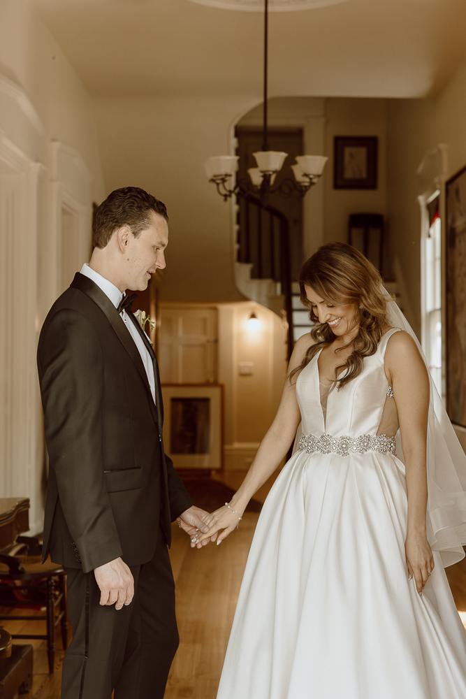 Bride and groom hold hands while both admire her wedding dress.