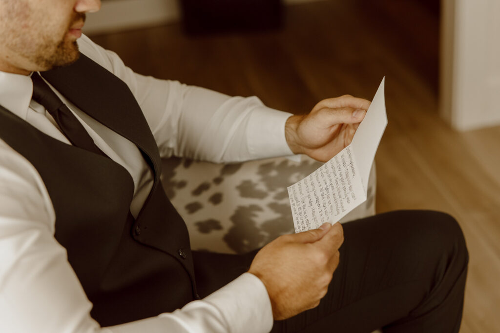 A groom sits on a chair and reads a handwritten letter from his bride.