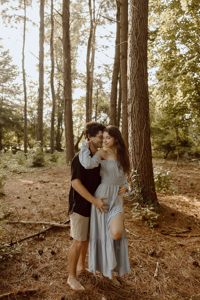 Couple stands close together while standing in the middle of the woods at sunset.