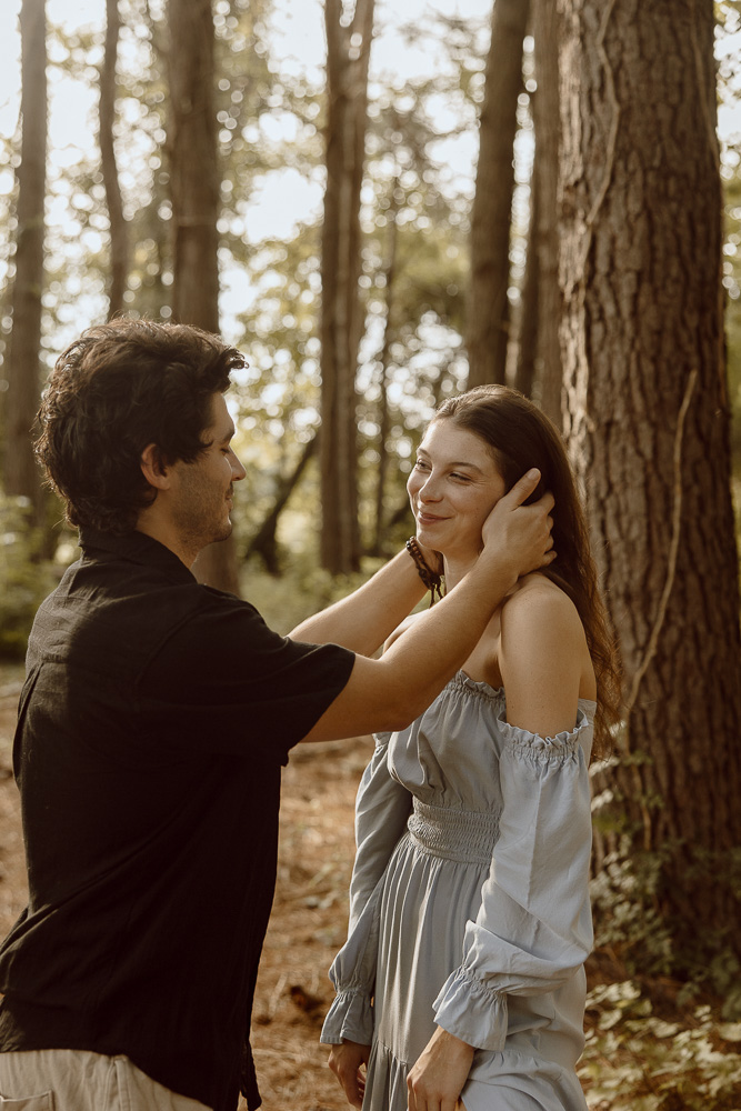 A couple stands together and smiles at each other while in the woods.