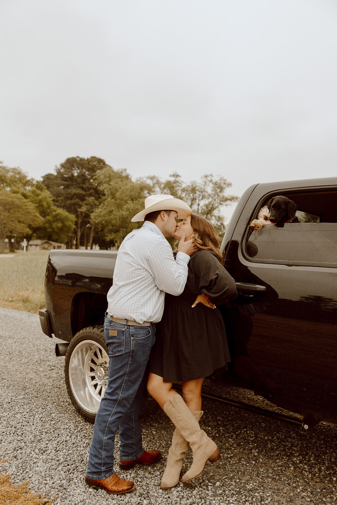 A couple shares a kiss while leaning against their truck while wearing cowboy boots.