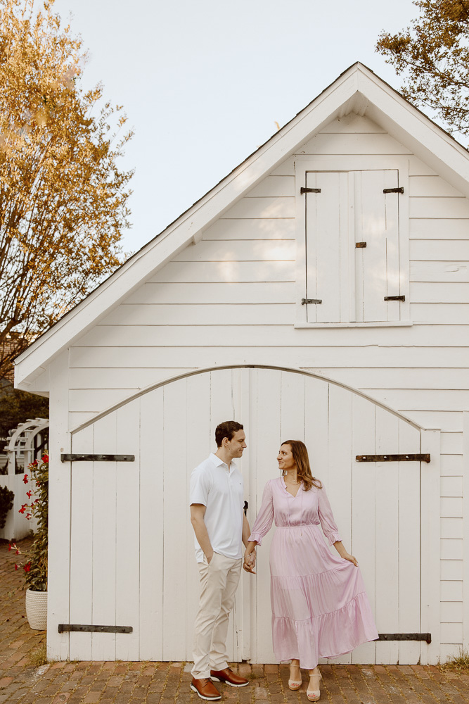 Engagement photos with couple standing in front of white barn and holds hands.