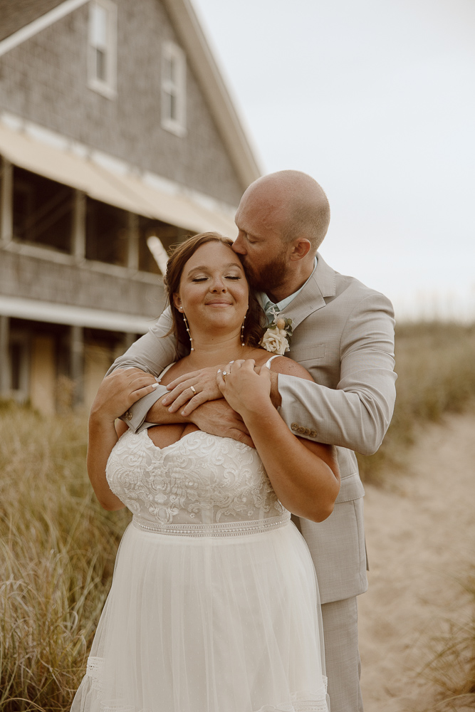 Wedding couple stands together and shares a kiss while at the beach.