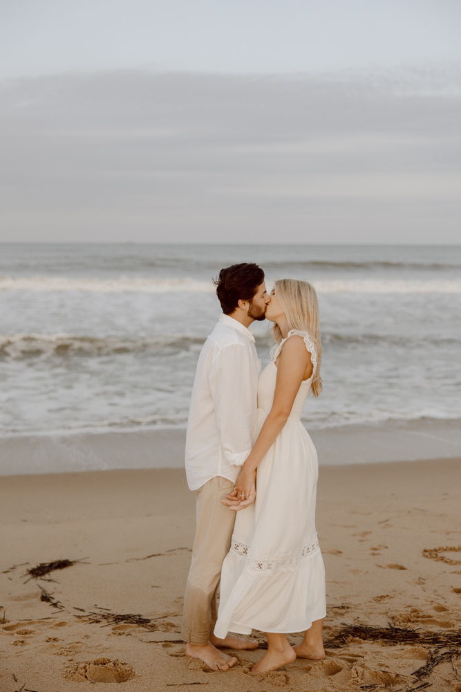 During their engagement session in Virginia Beach, a couple holds hands and shares a kiss with the waves crashing behind them.