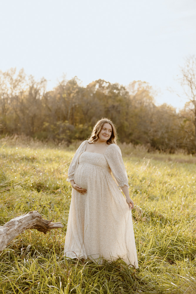 Women stands in grassy field during sunset, her dress flowing behind her and showing off her baby bump during a maternity session.
